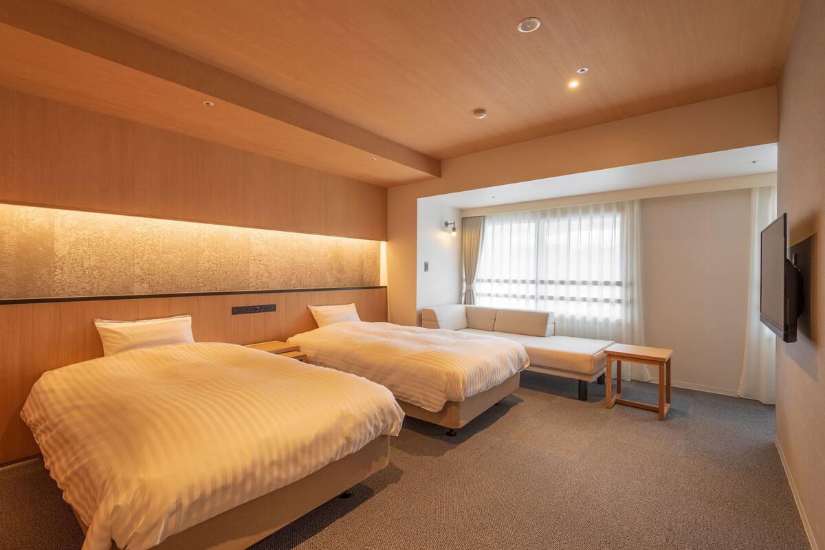A hotel room with two beds, a small table, a TV, and a window with curtains.