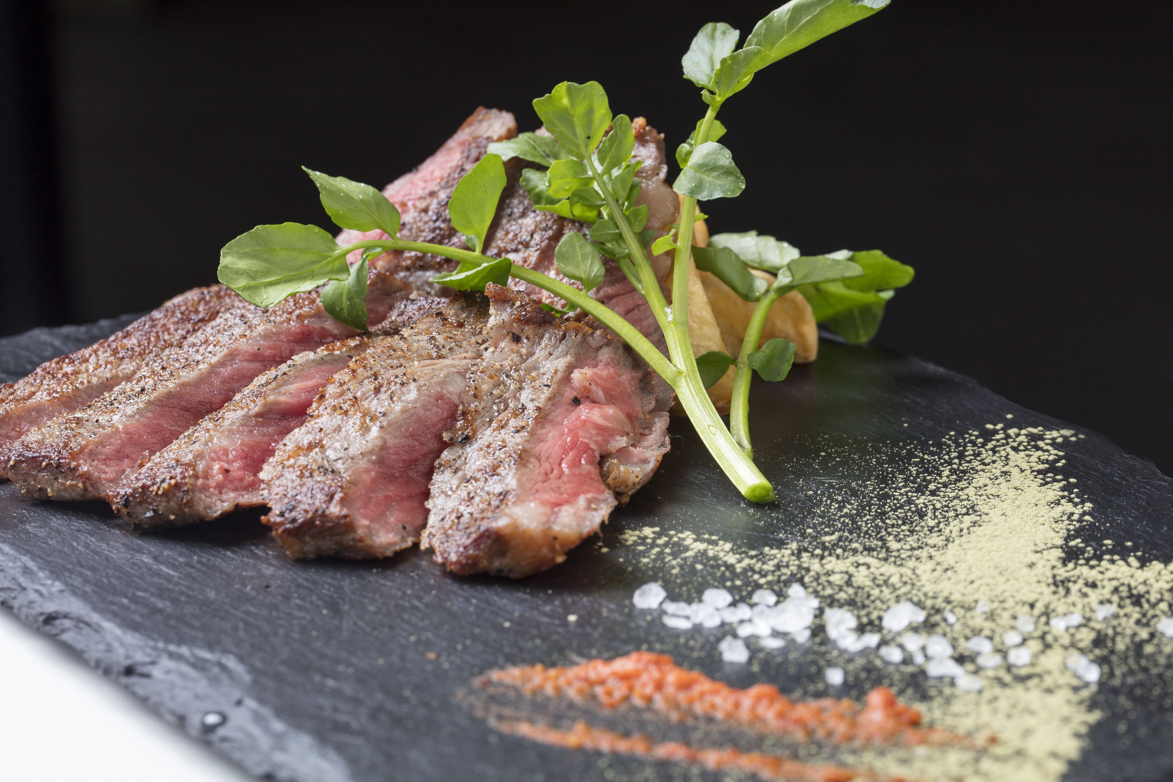 A steak with a garnish of green leaves and herbs, served on a slate plate with a sprinkle of seasoning.