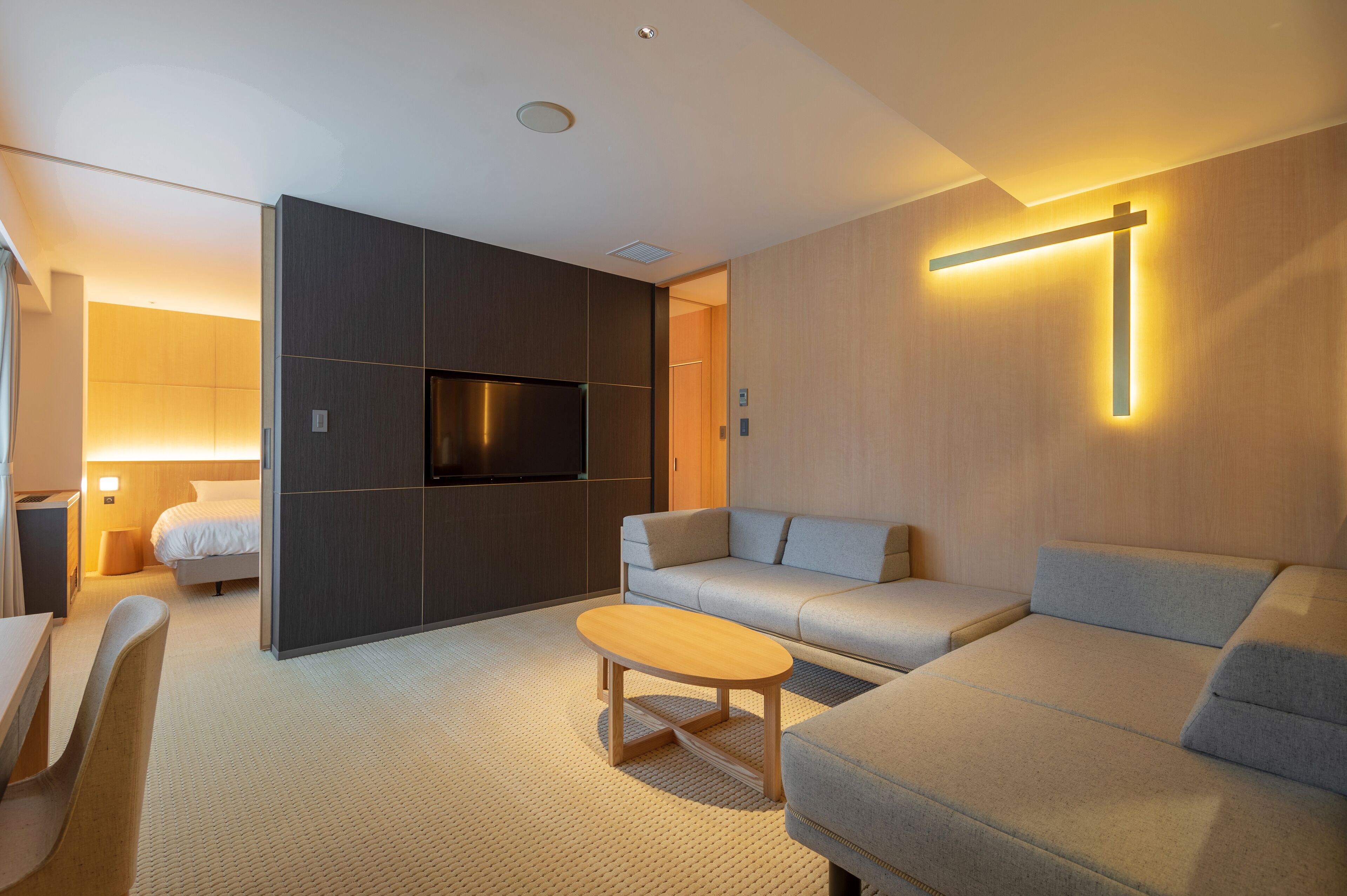 Una habitación de hotel moderna con sofá, una mesita de madera, televisión y una lámpara en forma de cruz en la pared.