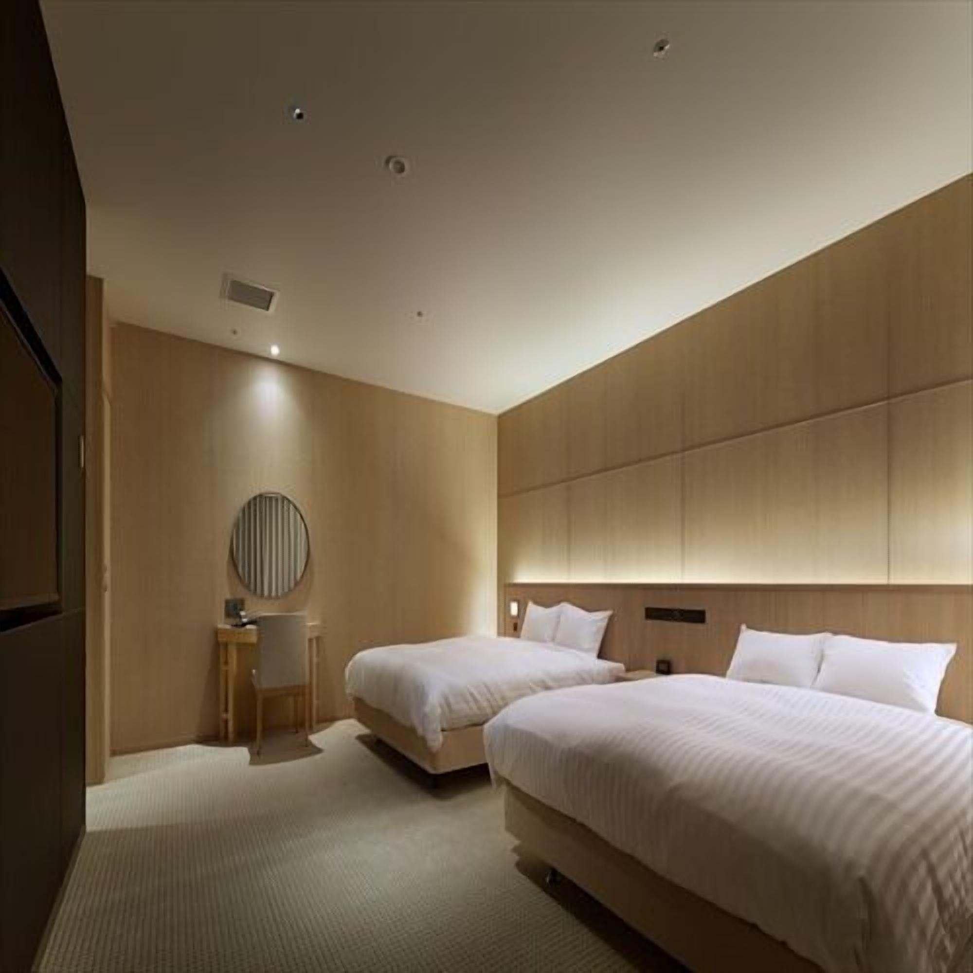 A modern hotel room with two beds, a round mirror, a small desk, and a built-in headboard.