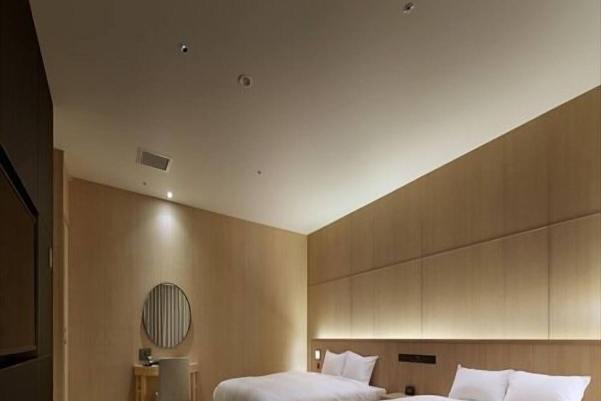 A modern hotel room with two beds, a round mirror, a small desk, and a built-in headboard.
