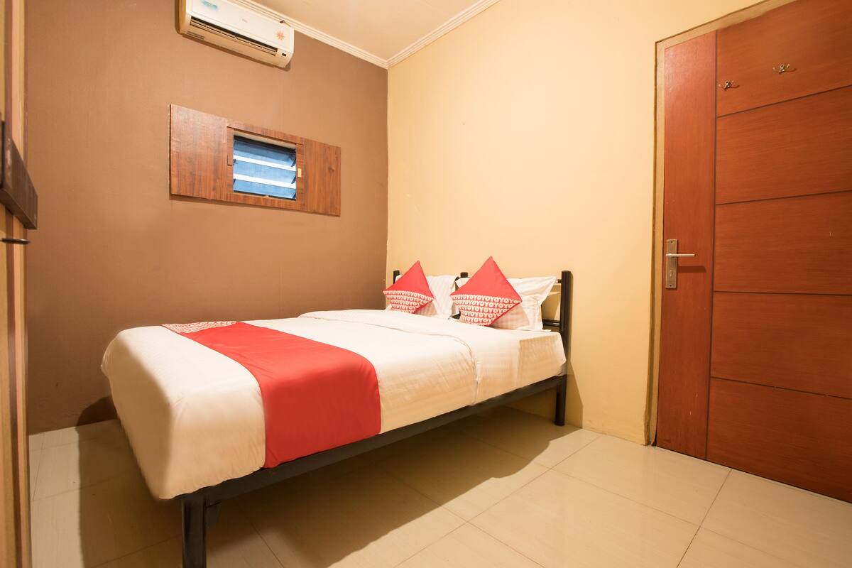 Una cama individual con una colcha roja y blanca, un ventanal pequeño con persianas y una puerta de madera.