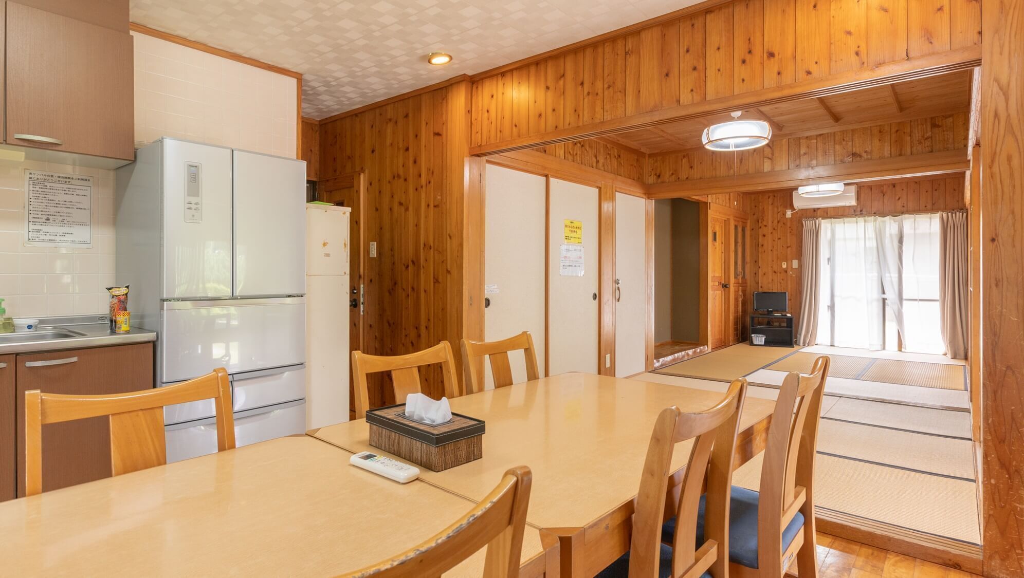 A traditional Japanese-style room with tatami flooring, a wooden dining table, chairs, a refrigerator, a television, and a sliding door leading to another room.