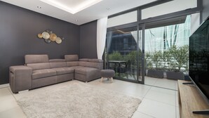 A modern living room with a sectional sofa, a small ottoman, a flat-screen TV, and a large sliding glass door leading to an outdoor area with plants.