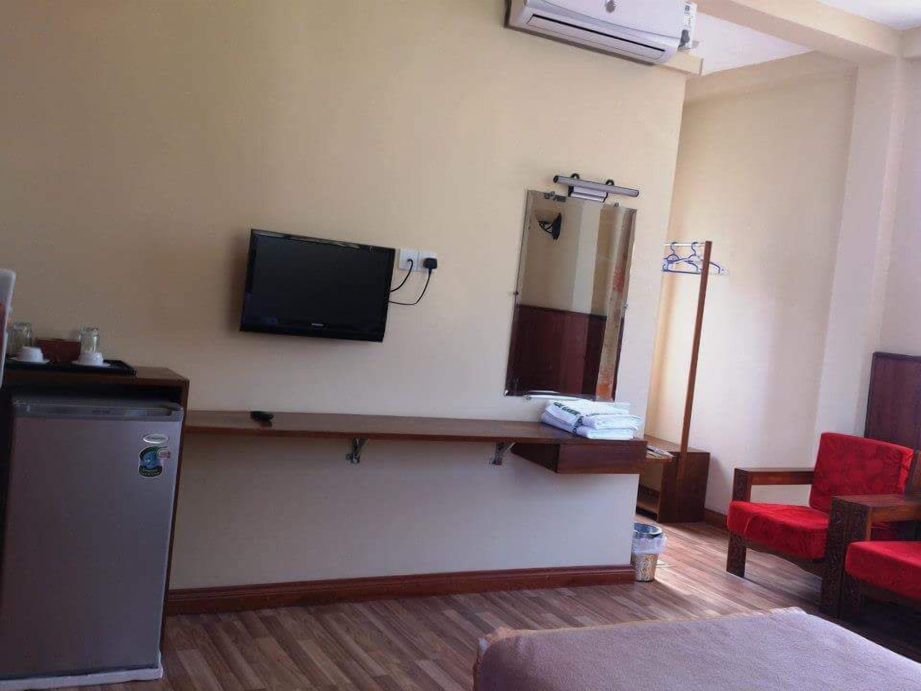 A hotel room with a bed, a desk with a computer, a television, a mirror, a red chair, and a small refrigerator.