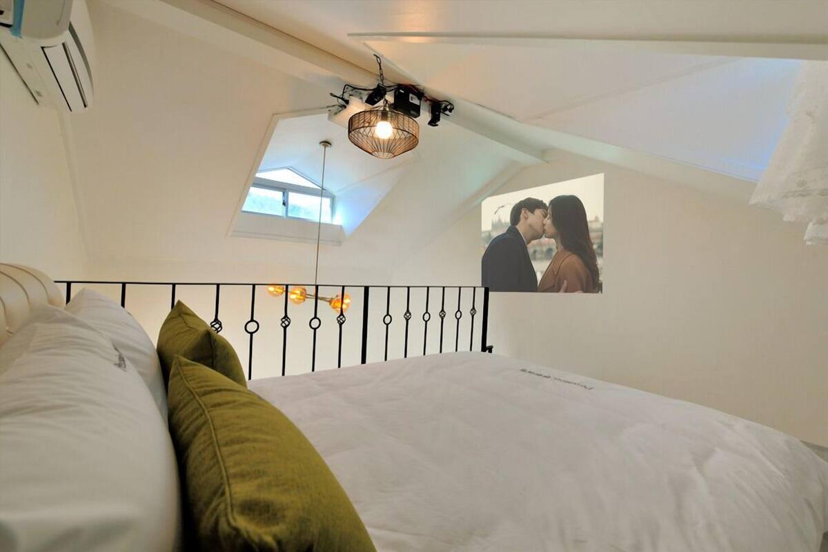 Un dormitorio con cama, lámpara colgante, ventana y espejo que refleja una pareja.