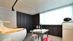 A compact room with a bathtub, a small dining table with two chairs, a refrigerator, and a window with blinds.