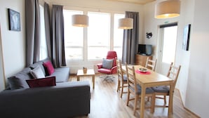 A modern living room with a sofa, red armchair, dining table, and chairs. There are large windows with curtains, a television, and a wooden ceiling. - Blue View Cabin 7A with Hot Tub (Bláskógabyggd)
