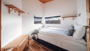 A small, well-lit bedroom with a single bed, a wooden bench, and two windows offering a view of a fence and sky. - Blue View Cabin 7A with Hot Tub (Bláskógabyggd)