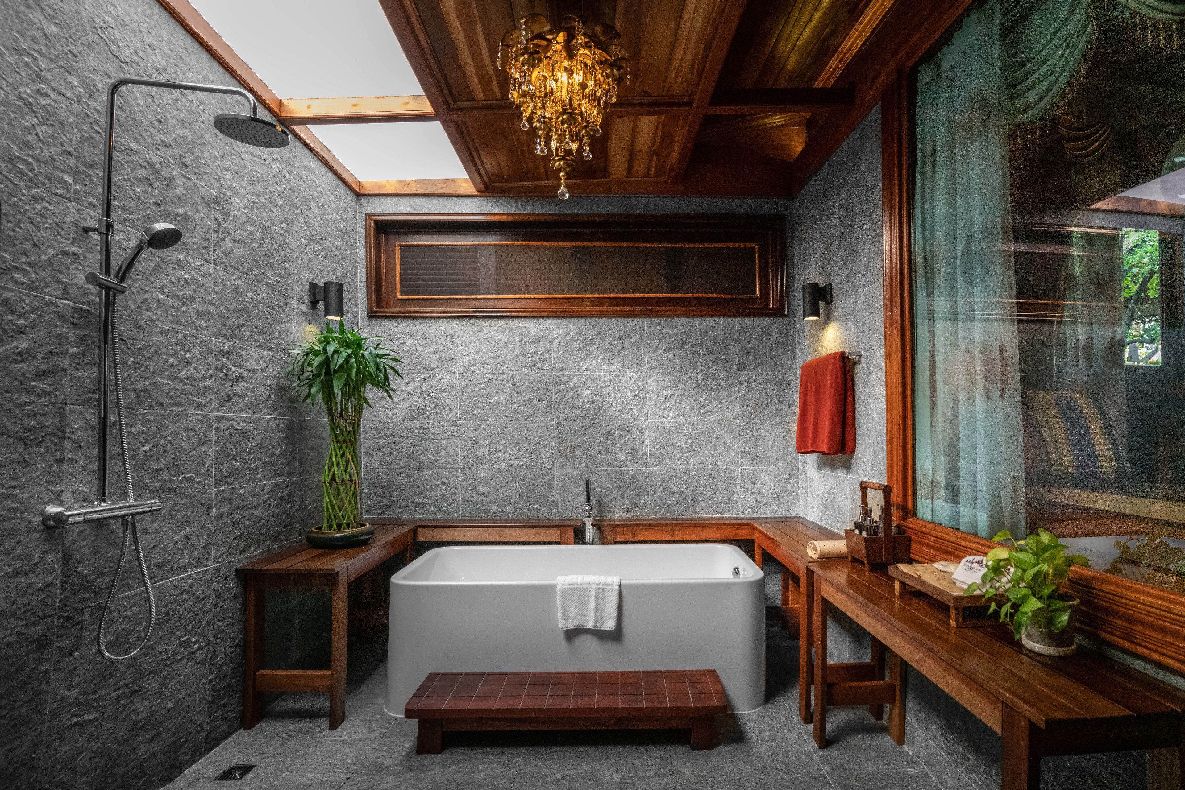 A modern bathroom with a freestanding bathtub, a shower, wooden benches, and a large window with a view of greenery.