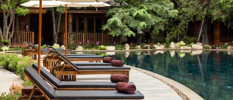 A poolside area with wooden lounge chairs and umbrellas, surrounded by lush greenery and a pool.