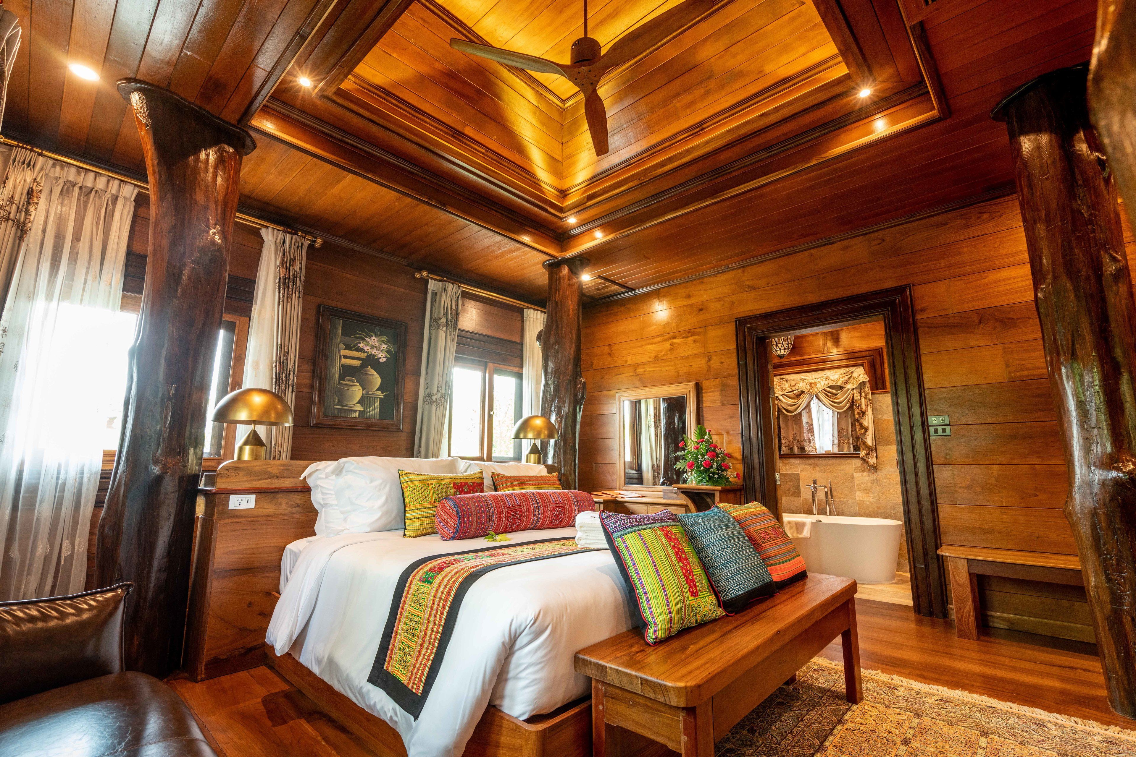 A spacious bedroom with a large bed, a wooden bench, a nightstand, a bedside table, a lamp, a window with curtains, and a bathroom with a bathtub and a mirror.