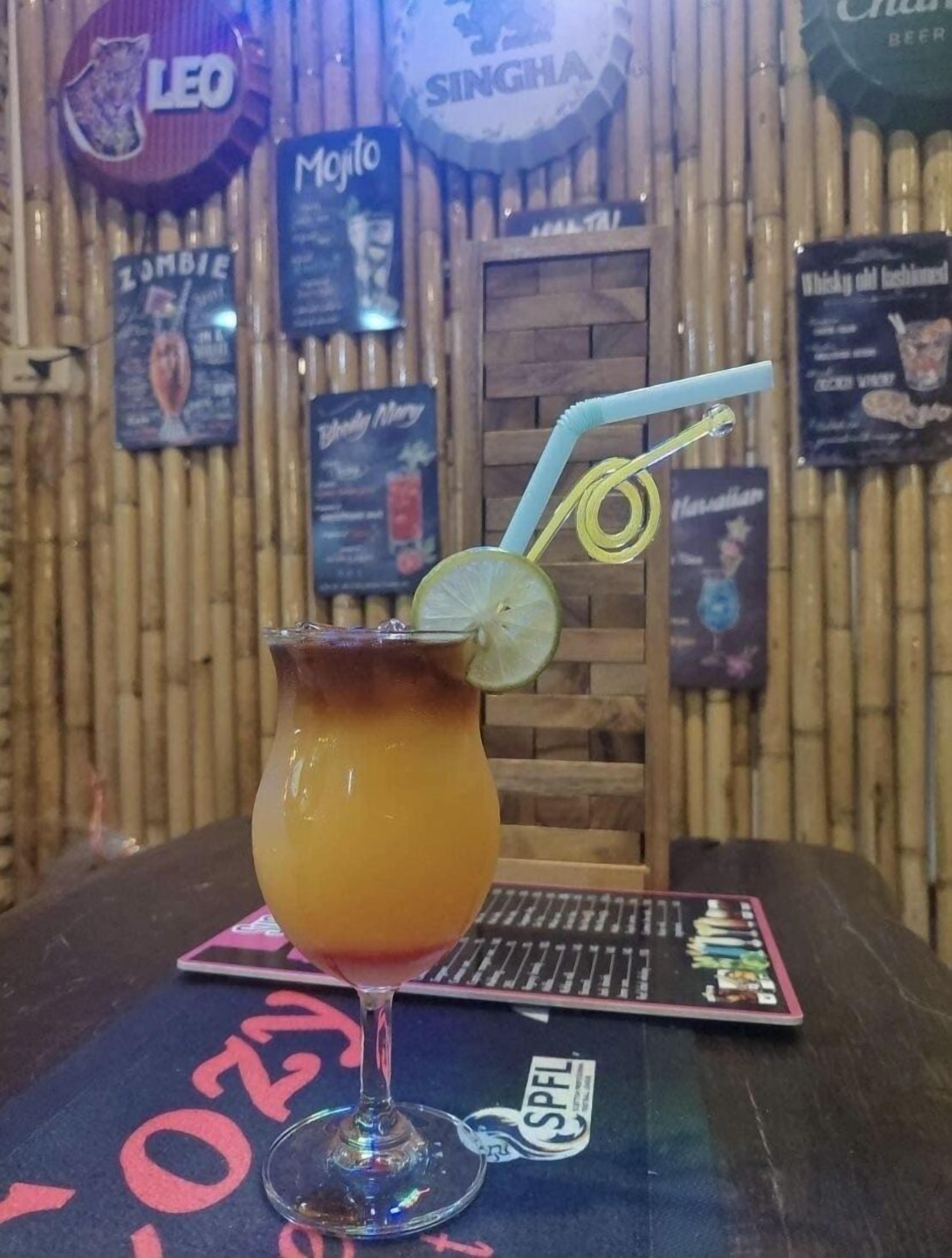 A cocktail in a glass with a lime wedge and a straw, placed on a table with a card game and a menu board in the background.