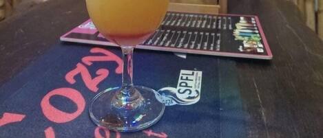 A cocktail in a glass with a lime wedge and a straw, placed on a table with a card game and a menu board in the background.