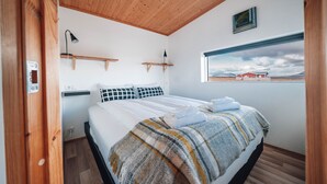 Quarto com cama, teto de madeira, paredes brancas, janela com vista e uma pequena estante com abajur.