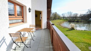Un balcón con mesa y sillas de madera, vista a un lago, y un edificio blanco con una ventana y una puerta.