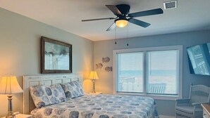 Um quarto com cama, duas luminárias de cabeceira, ventoinha no teto, janela com persianas, uma cadeira e cômoda.