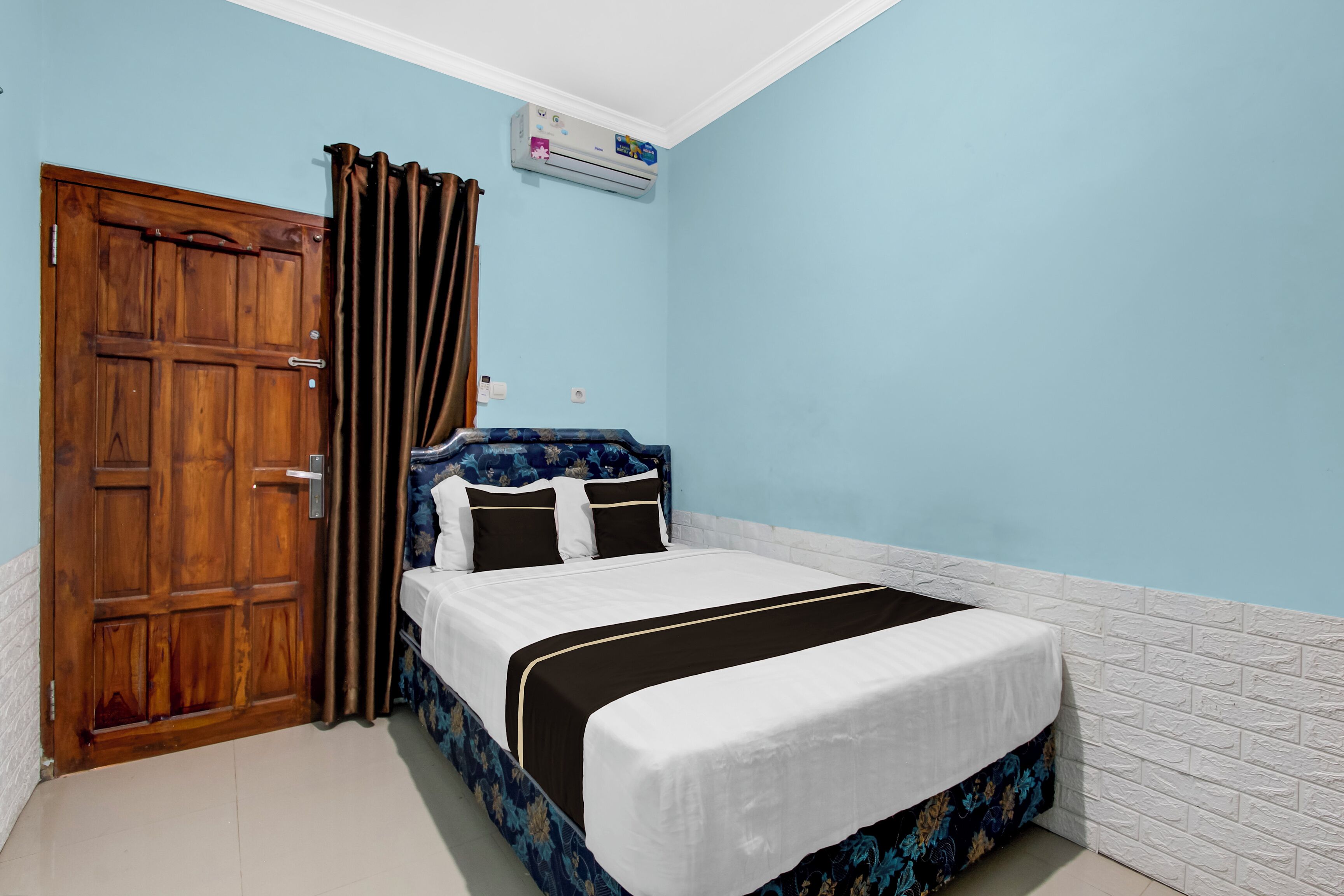 A bedroom with a wooden door, a bed with a floral headboard, and an air conditioning unit on the wall.