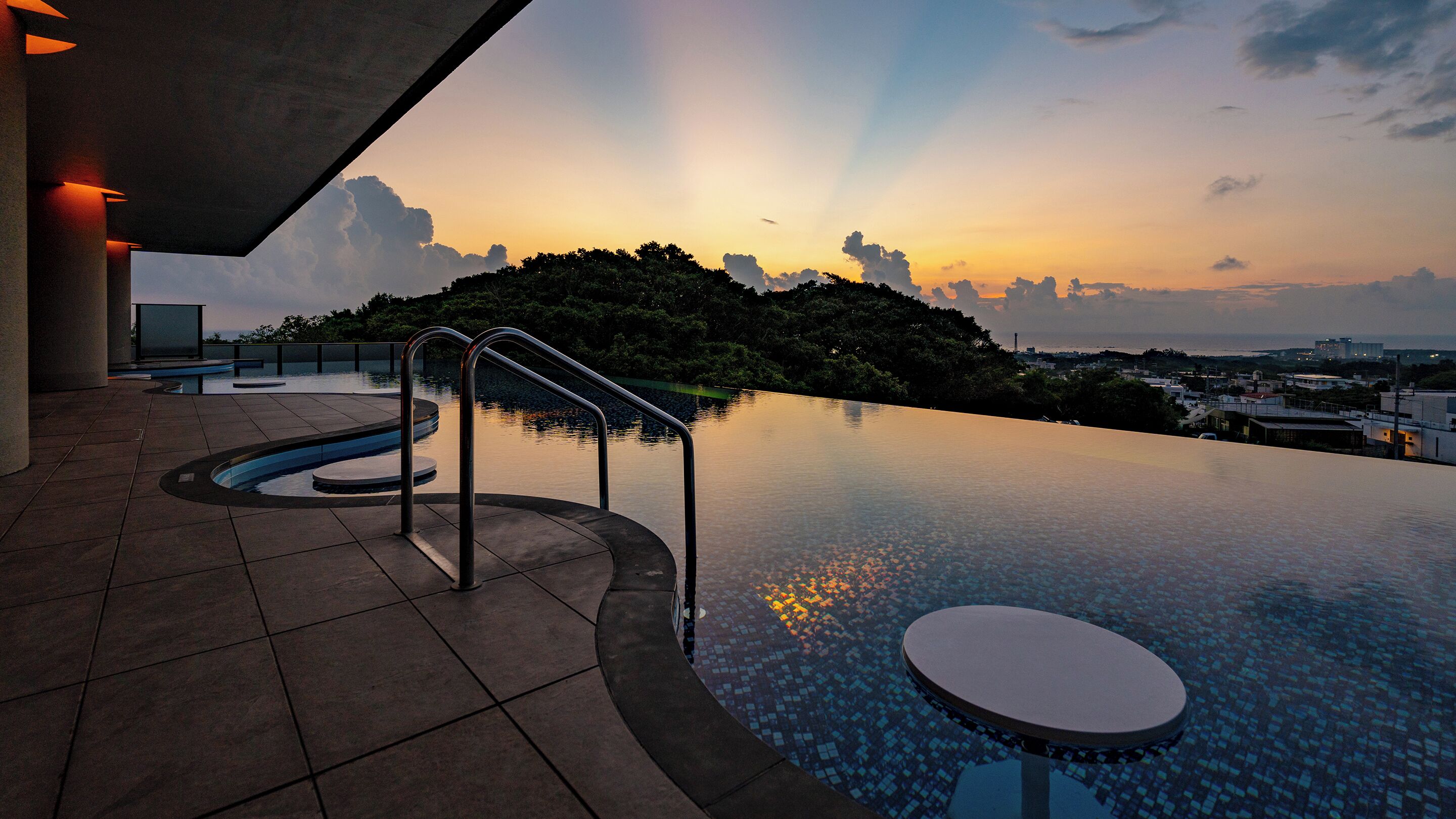 Uma piscina infinita com vista para um pôr do sol sobre uma colina, rodeada por um edifício moderno com piso em azulejos e guarda-corpos de metal.