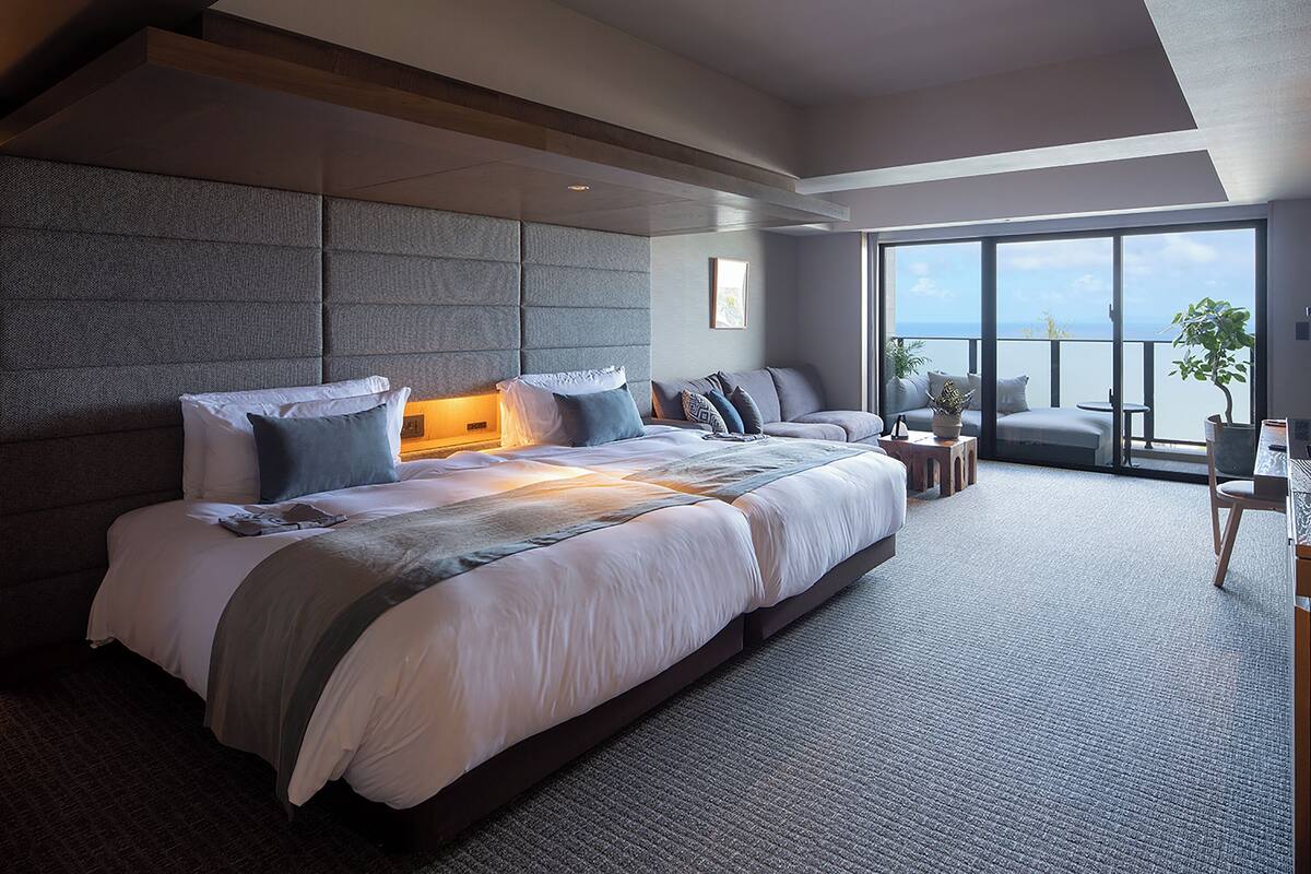 A modern hotel room with a large bed, a seating area with a view of the sea, and a balcony with a potted plant.