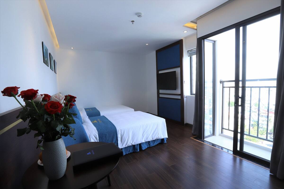 A modern hotel room with a bed, a desk, a vase with flowers, and a large window with a balcony.