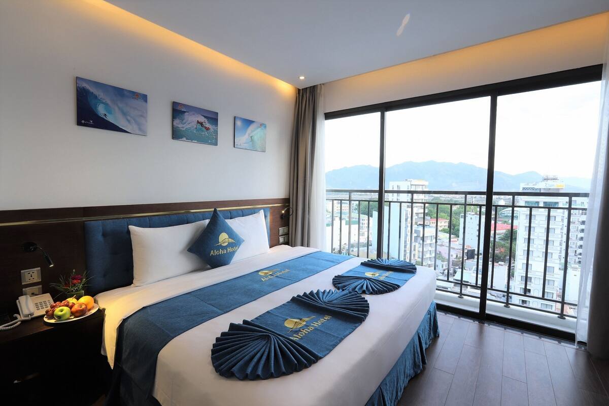 A hotel room with a large bed, a nightstand with a phone, and a view of the city through large windows.