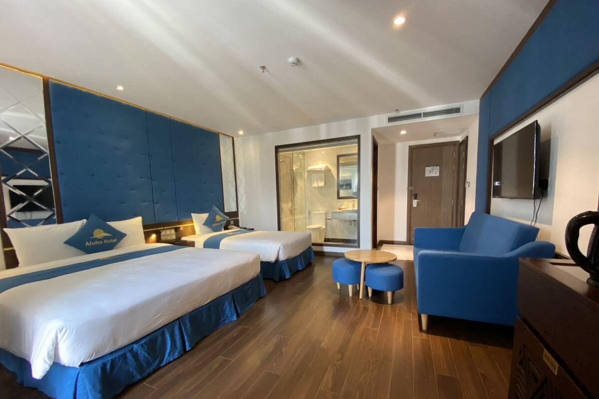 A hotel room with two beds, a seating area with a blue chair and ottoman, a TV, and a bathroom visible through a glass door.