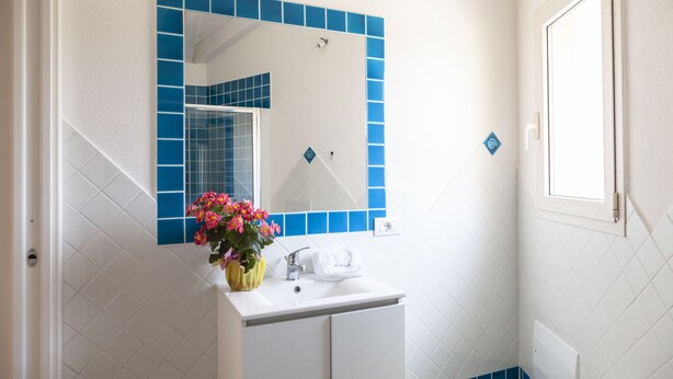 Une salle de bain avec un lavabo blanc, des murs recouverts de carreaux bleus, un miroir et une fenêtre.