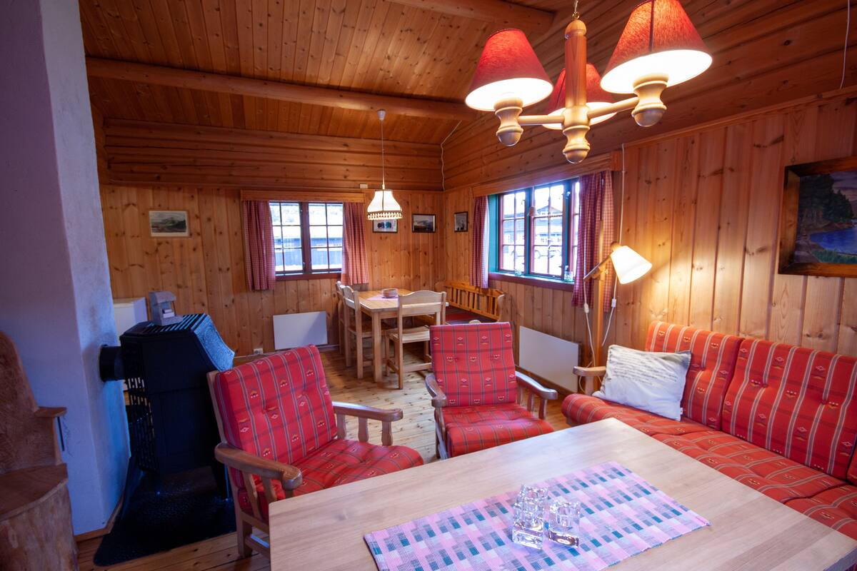 A wooden cabin interior with a dining area, red upholstered chairs, a sofa, and a table with glasses and a placemat.
