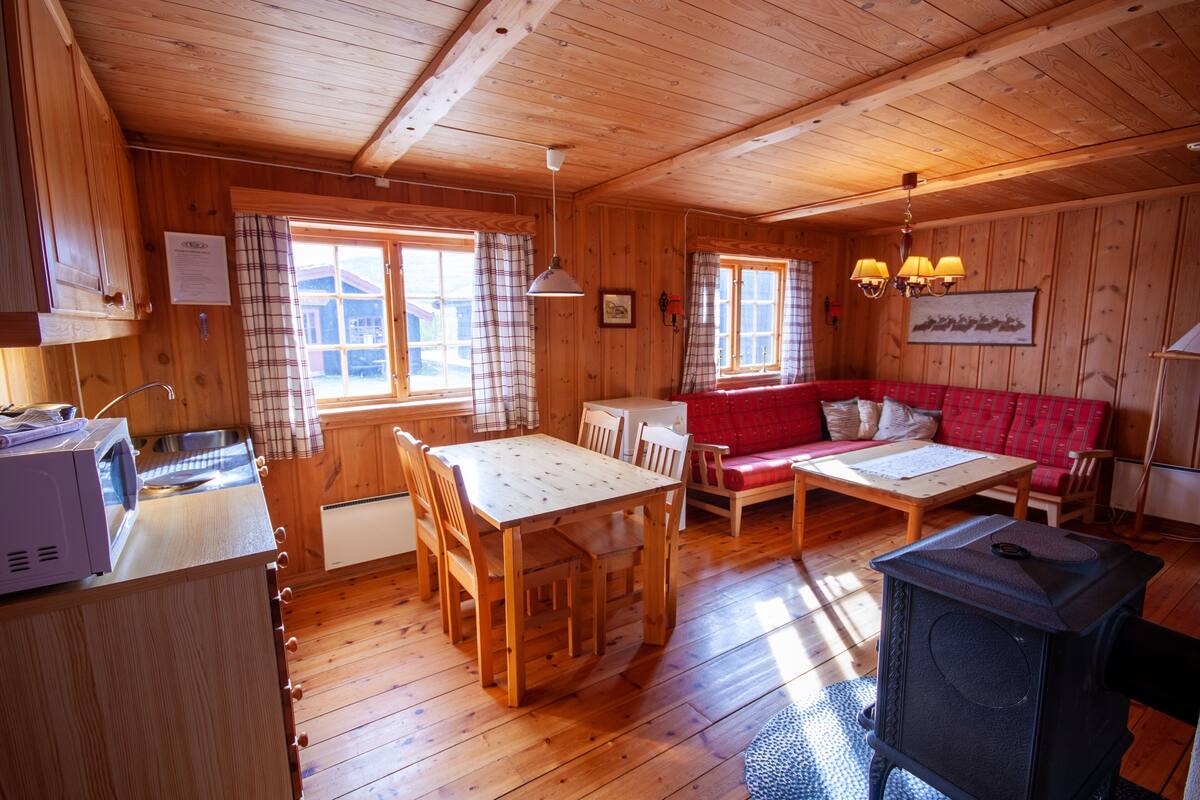 A wooden cabin interior with a dining table, red sofa, and a wood-burning stove.
