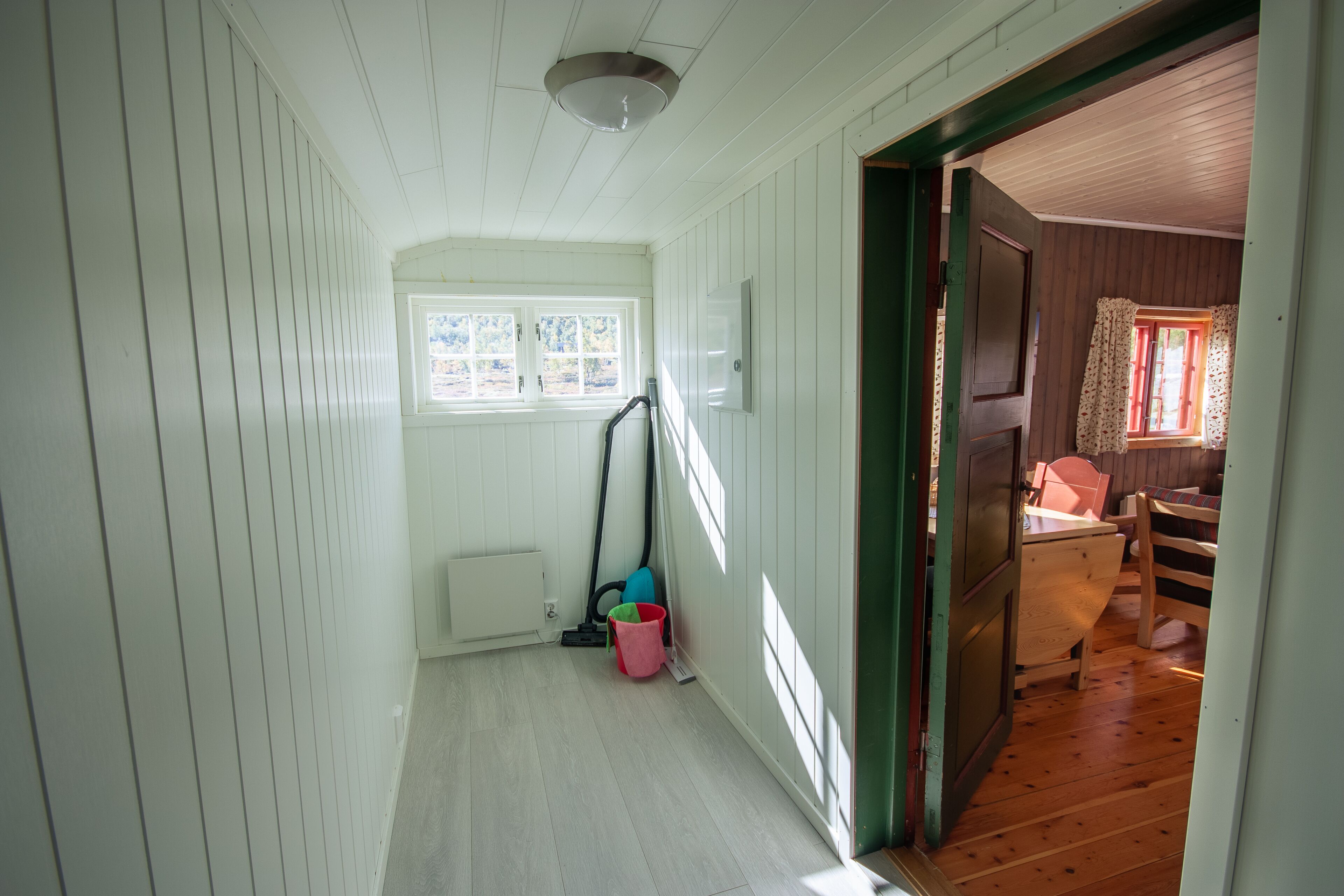 Un pasillo estrecho con paredes blancas y piso de madera, que conduce a una habitación con ventana y puerta.