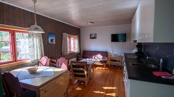 A cozy cabin-style room with a wooden floor, a dining table with chairs, a sofa, a kitchen area with a sink, and a television mounted on the wall.