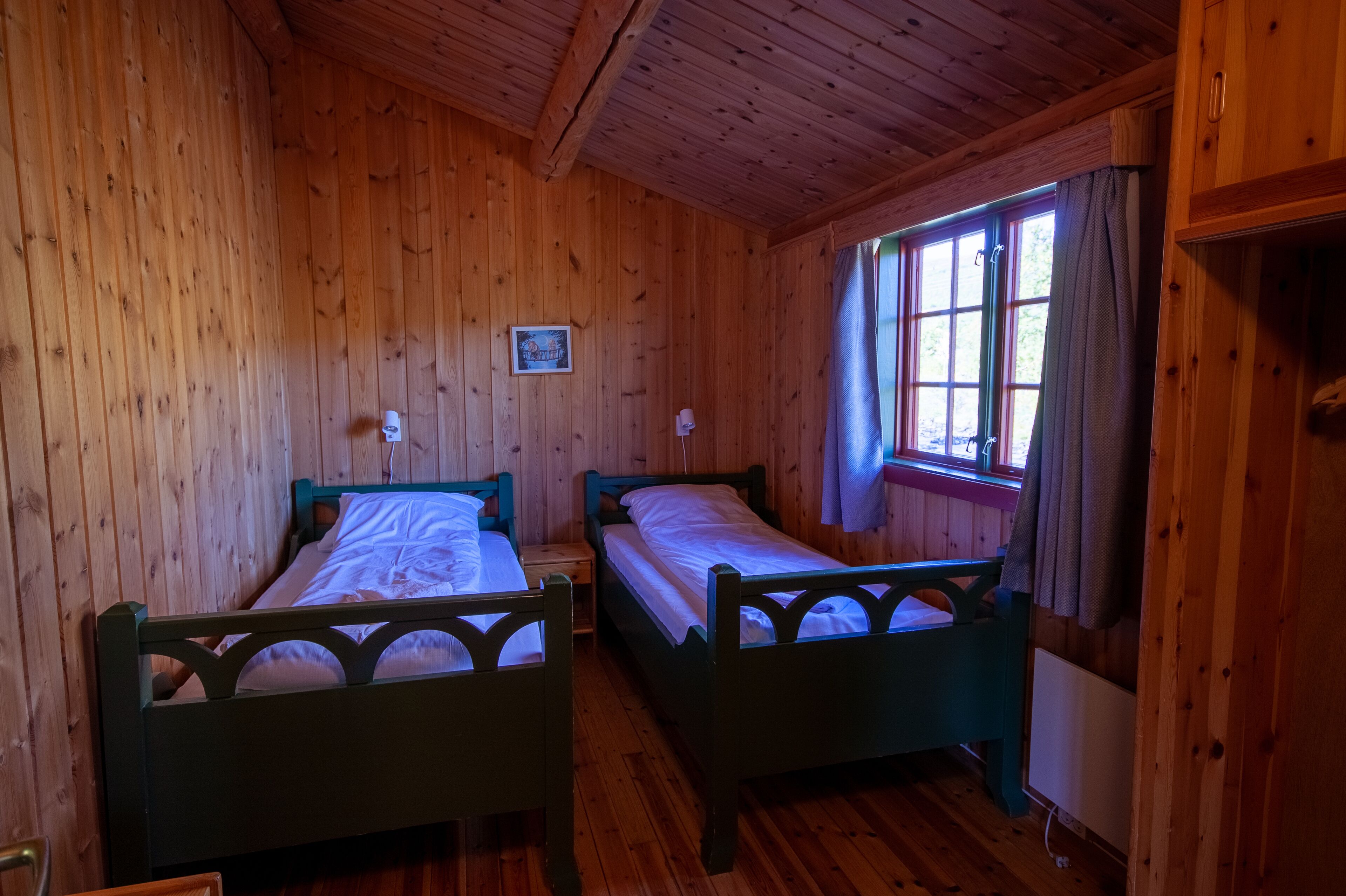 Ein Holzhütten-Zimmer mit zwei Betten, Holzwänden, einem Fenster mit Vorhängen und einem kleinen Bild an der Wand.