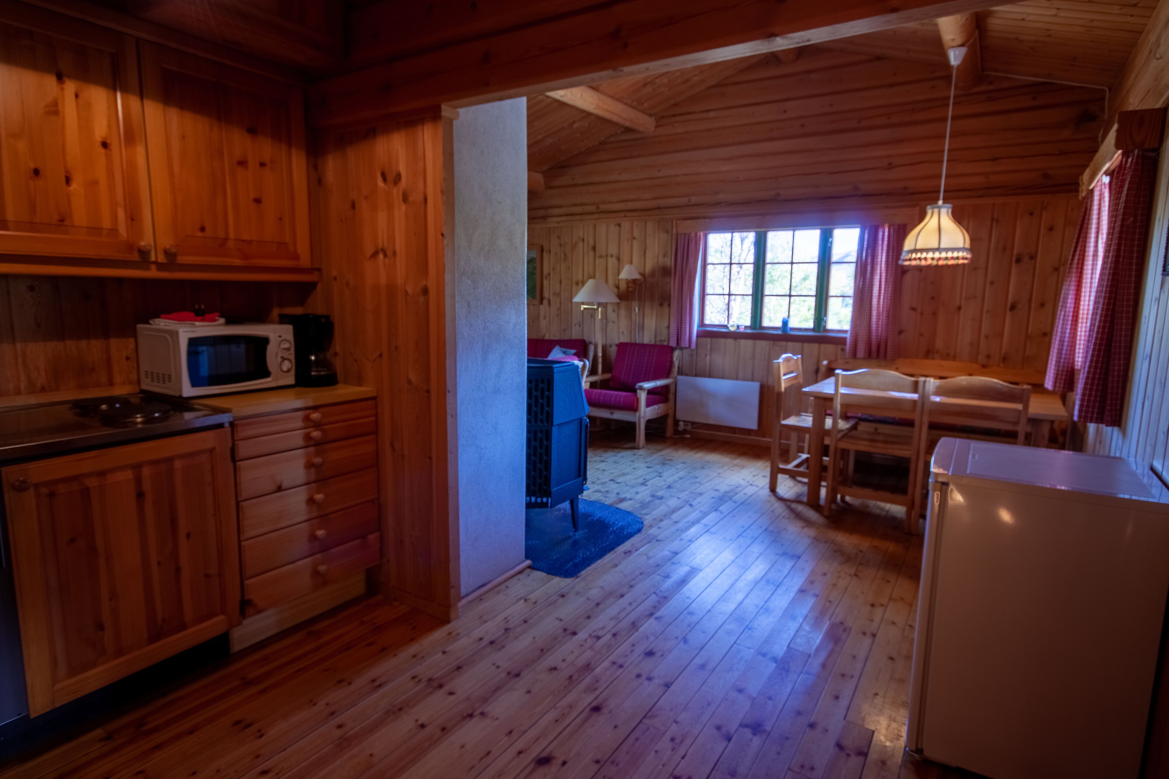 A wooden cabin interior with a kitchen, dining area, and a window with curtains.