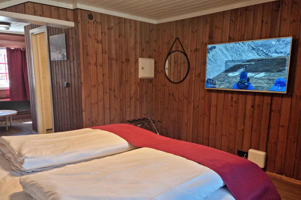 A hotel room with two beds, a wooden wall, a TV displaying a snowy landscape, and a red blanket on one bed.