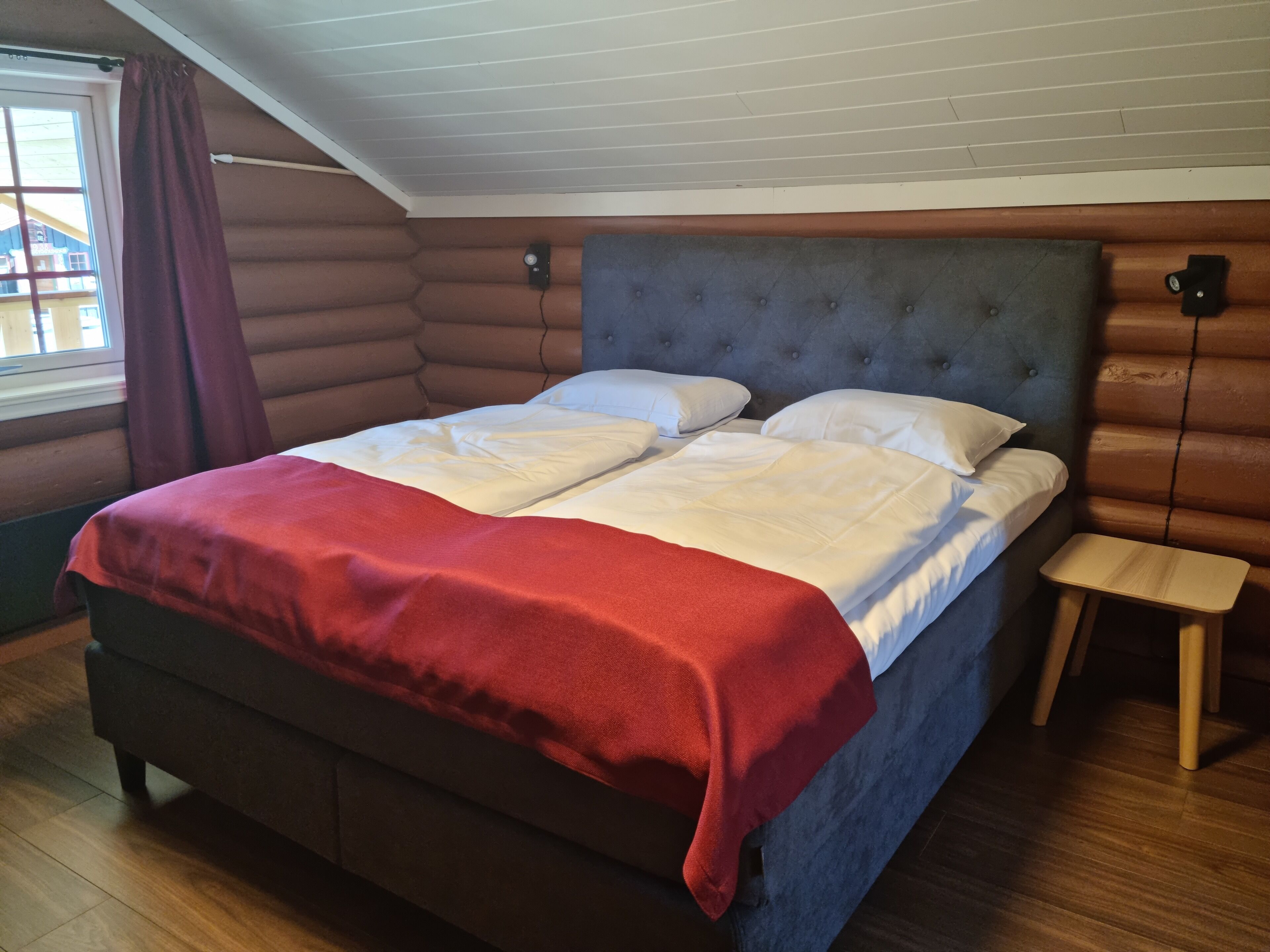 A bed with a red blanket, a wooden nightstand, and a window with curtains.