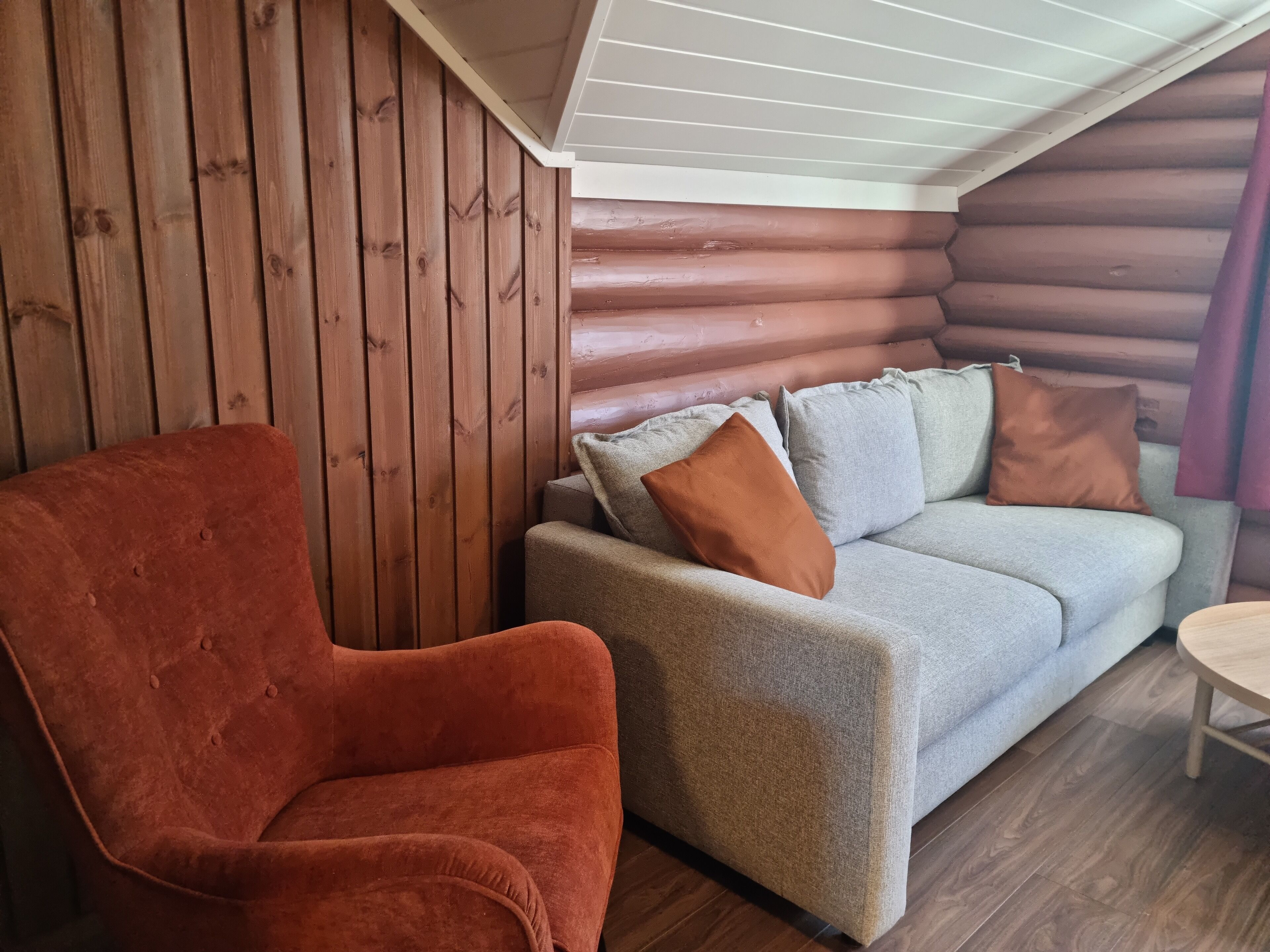 A cozy log cabin interior with a wooden wall, a grey sofa with cushions, and a red armchair.