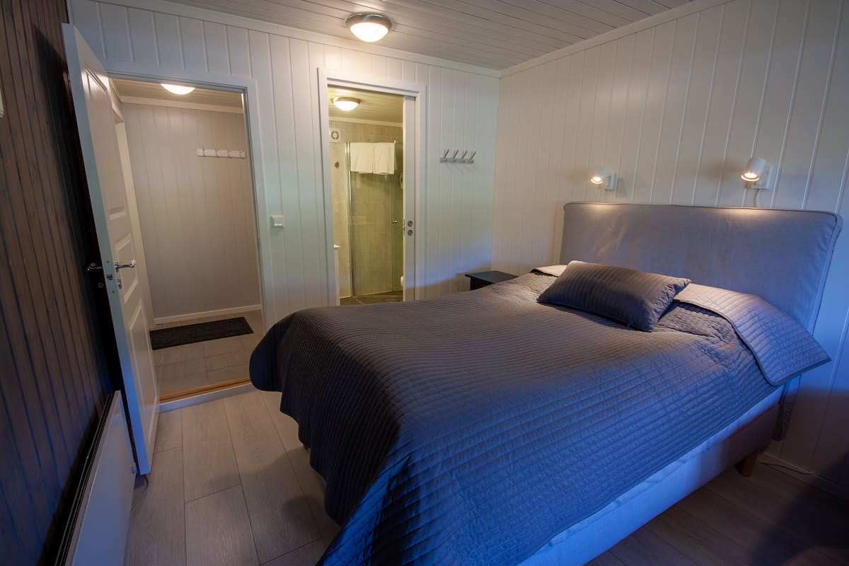 A bedroom with a bed, a nightstand, a door leading to a bathroom, and a wall-mounted light fixture.