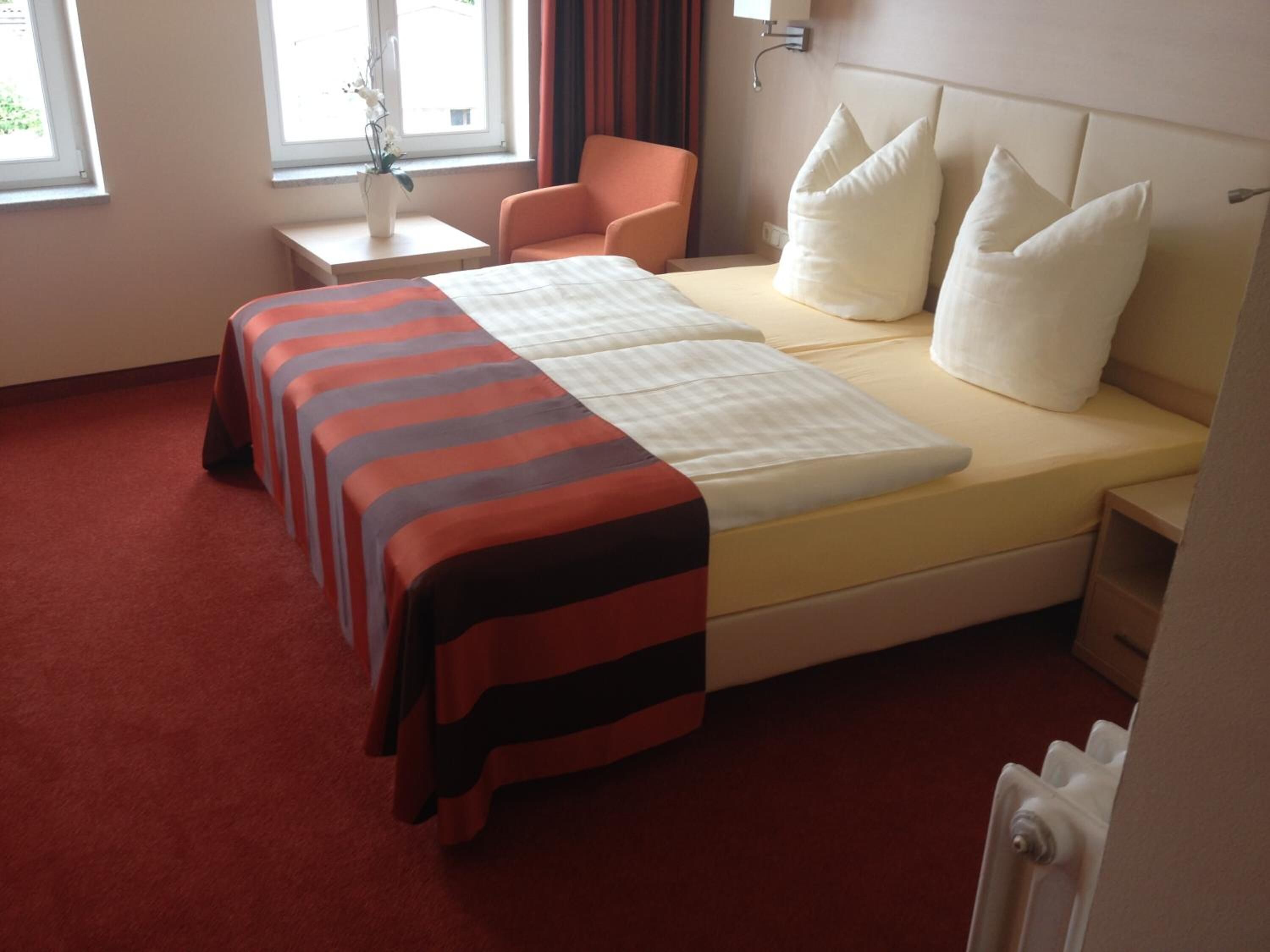 Ein Hotelzimmer mit Doppelbett, einem Sessel, einem Nachttisch und einem Fenster mit Vorhängen.