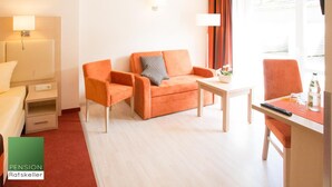 Une chambre d’hôtel moderne, équipée d’un canapé, d’un fauteuil et d’une petite table. On y trouve une fenêtre avec des rideaux, une table de chevet avec une lampe et un lit doté d’une tête de lit blanche.