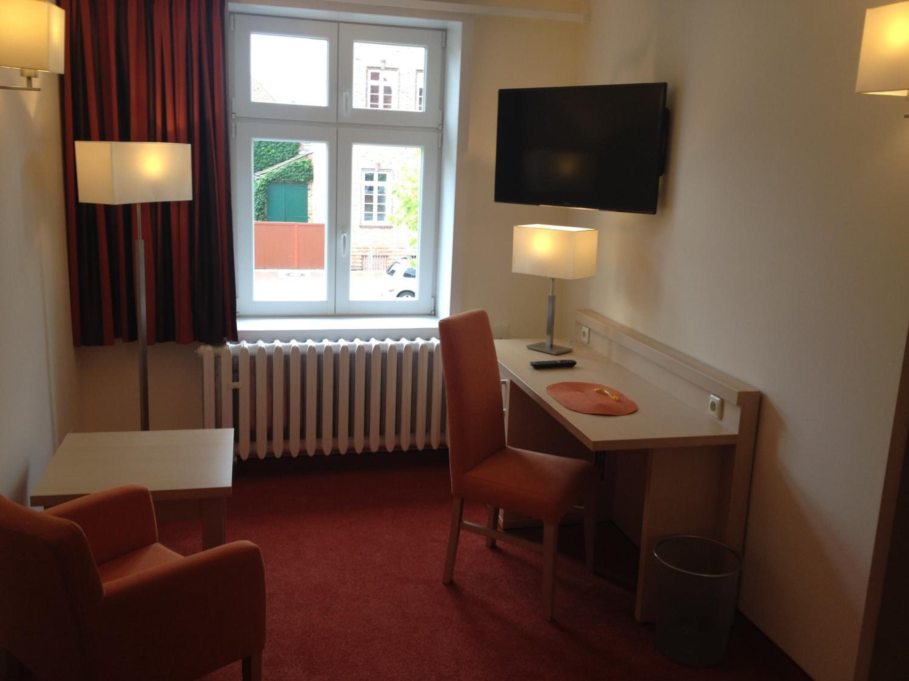 A compact hotel room with a single bed, a desk with a chair, a television, and a window with curtains.