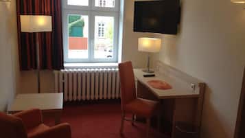 A compact hotel room with a single bed, a desk with a chair, a television, and a window with curtains.