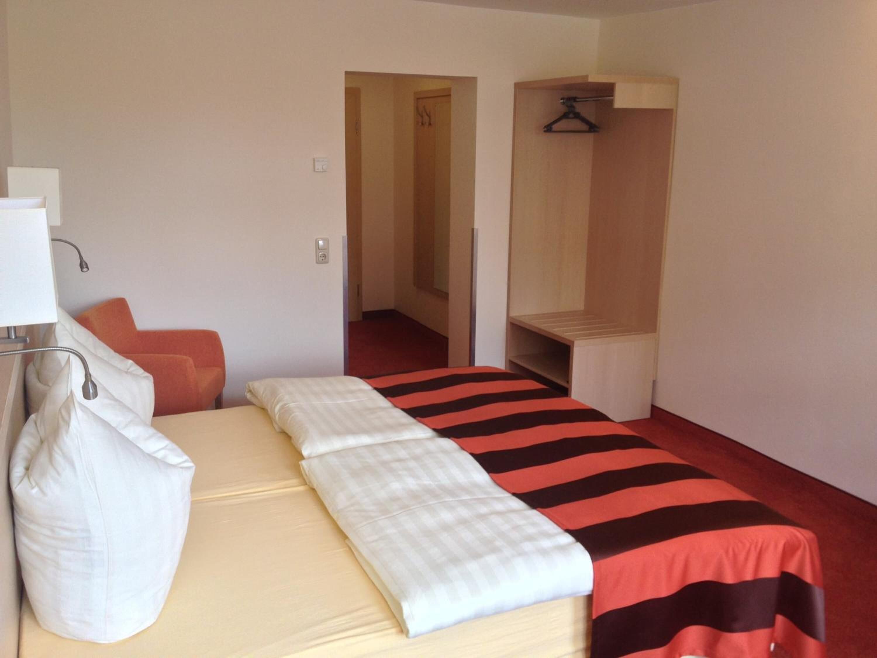 A single-bed room with a striped bedspread, a bedside table, a hanging coat rack, and a small seating area with a chair and cushions.