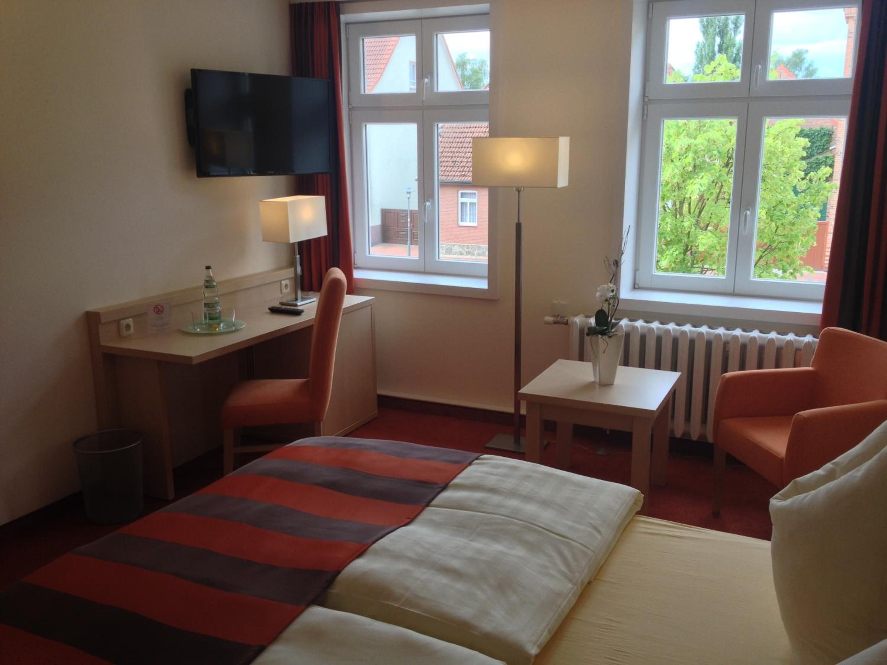 A hotel room with a bed, desk, chair, lamp, and windows.
