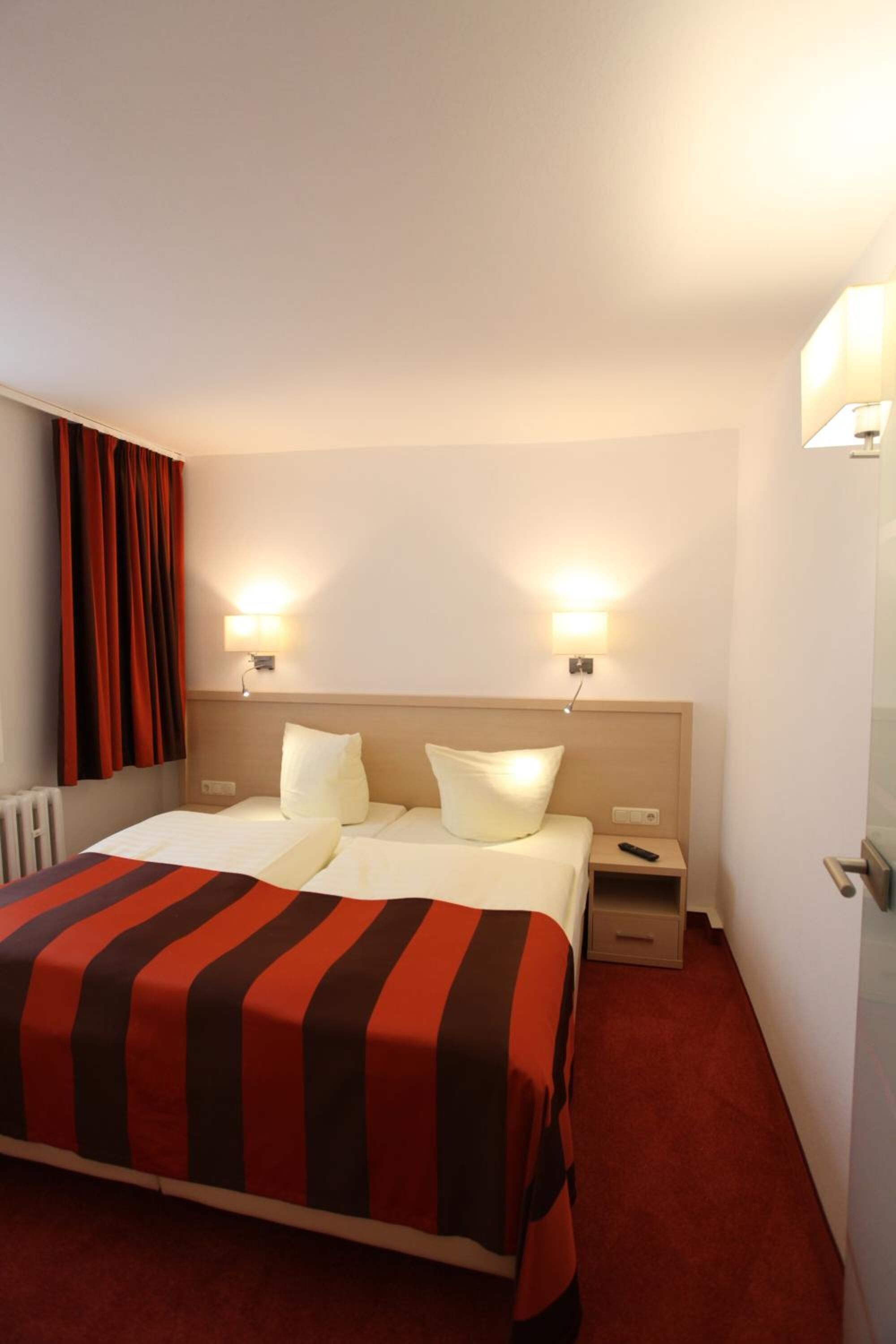 Habitación de hotel con una cama, dos lámparas de noche, una mesita de noche y una alfombra roja.