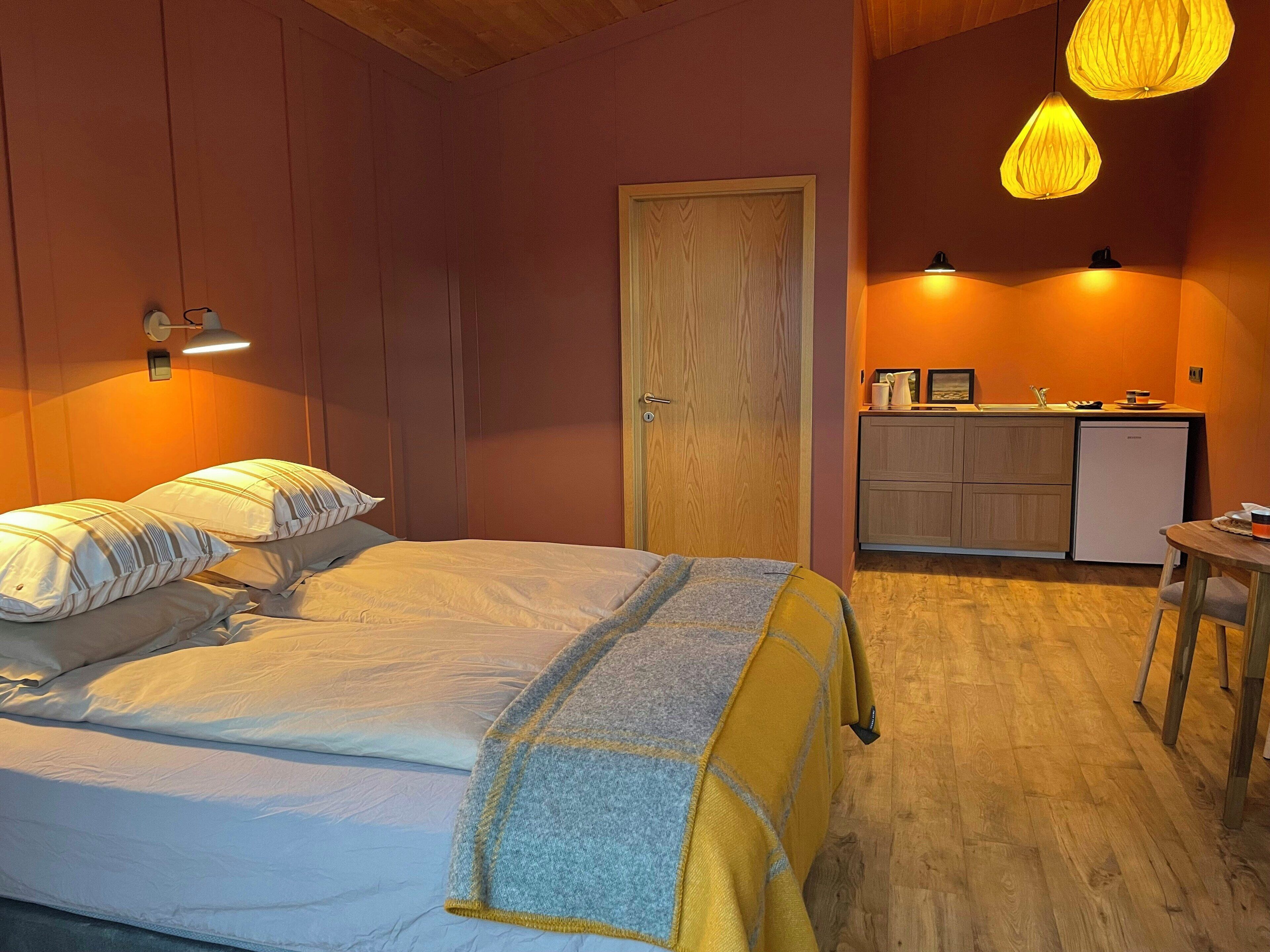 Un dormitorio con cama, lámparas de noche, una puerta de madera, una kitchenette y una pequeña mesa.