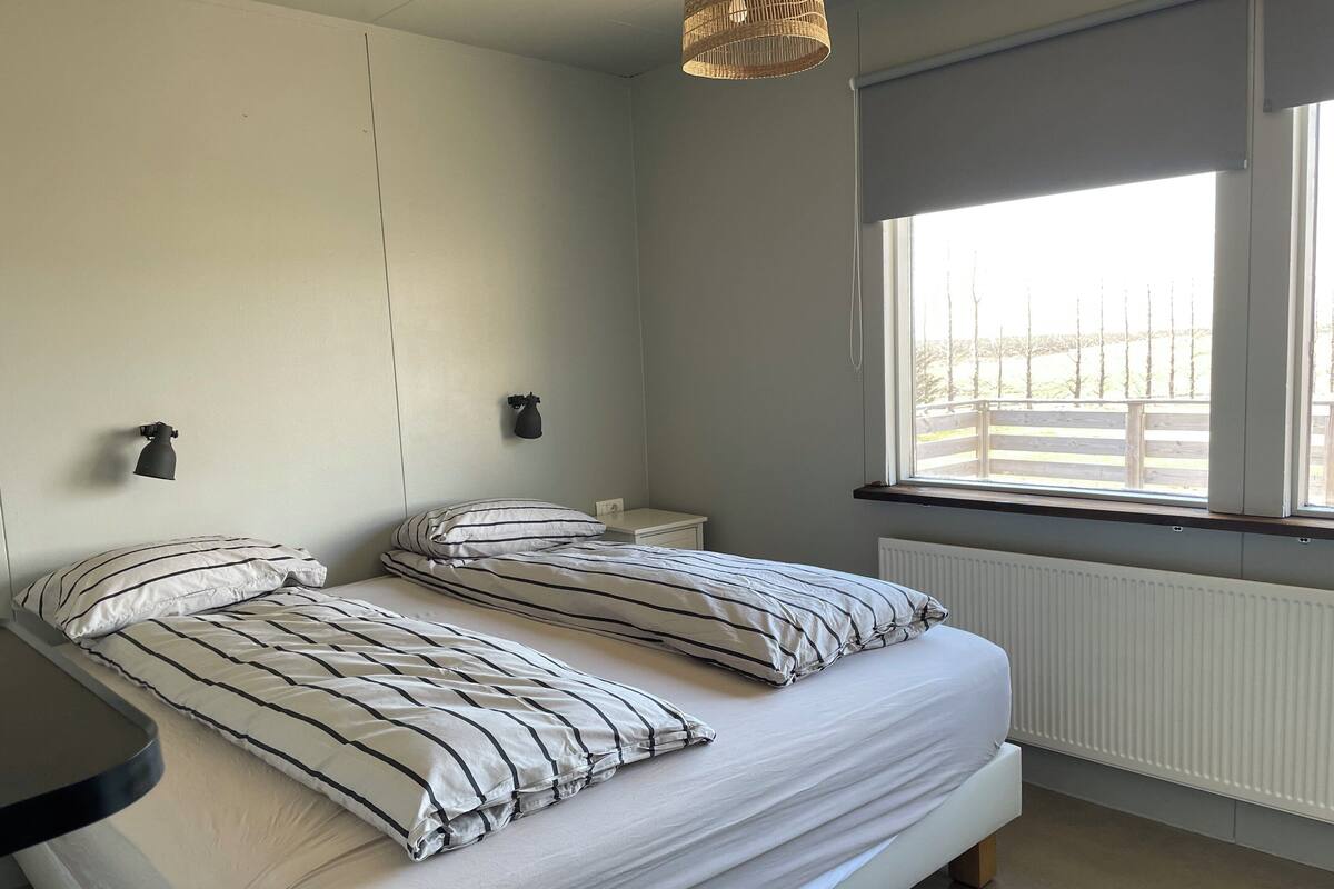 Une chambre avec deux lits, une fenêtre donnant sur une terrasse en bois, une lampe fixée au mur et un radiateur.