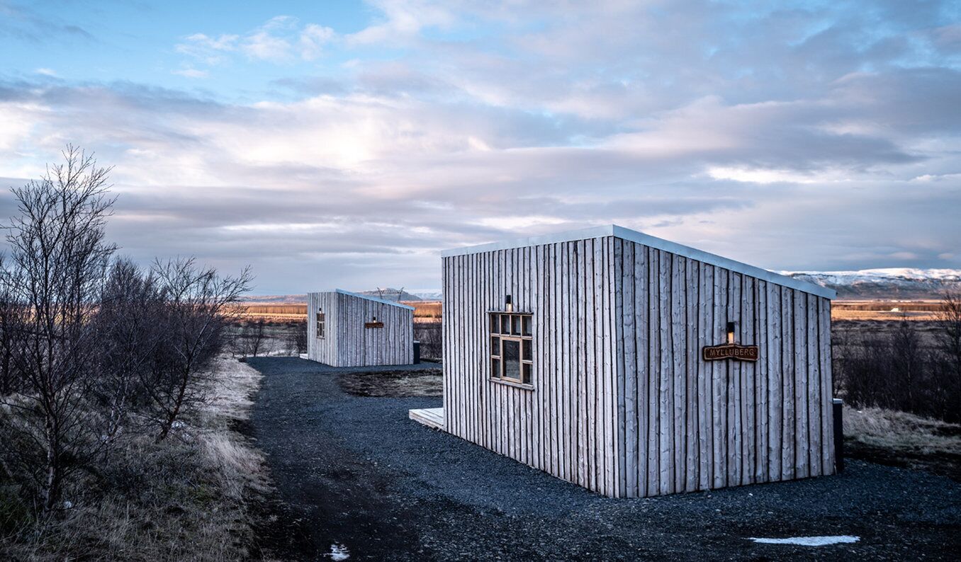A row of wooden cabins with a gravel path leading to them, set against a backdrop of a cloudy sky and distant mountains.