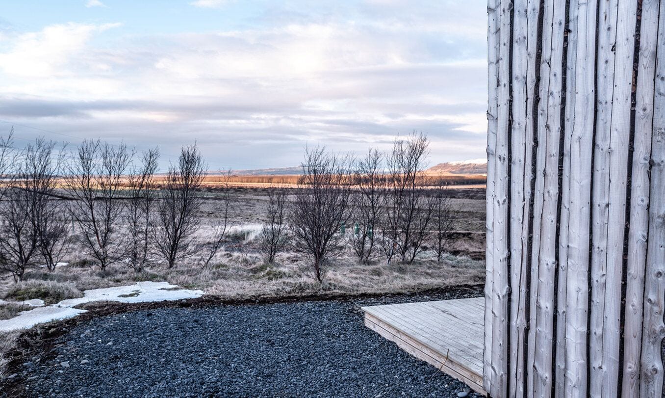 Eine Holzterrasse führt zu einem Gebäude mit Blick auf eine karge Landschaft, vereinzelte Bäume und ferne Berge.