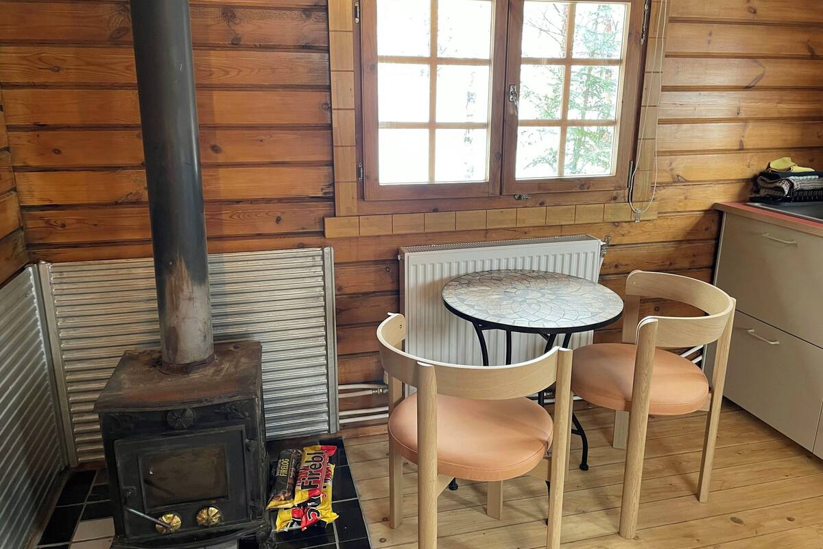 L’intérieur d’un chalet en bois, avec un poêle à bois, une table avec des chaises, une fenêtre et un radiateur.